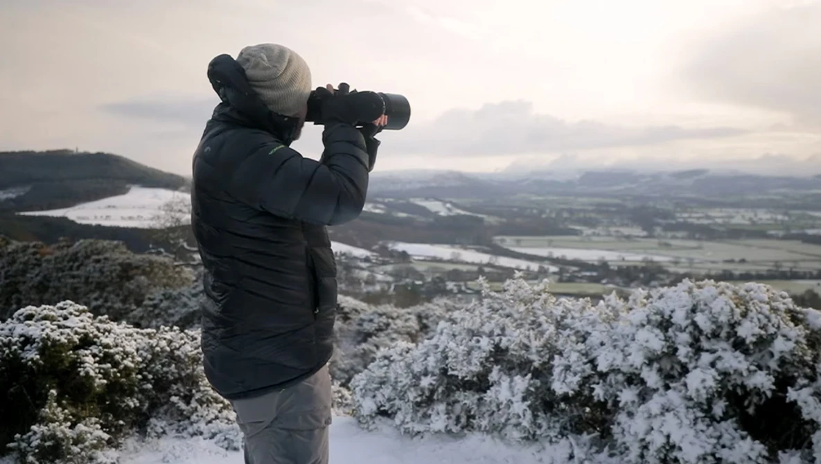 Photographer with telephoto lens capturing a snowy landscape from an elevated vantage point.
