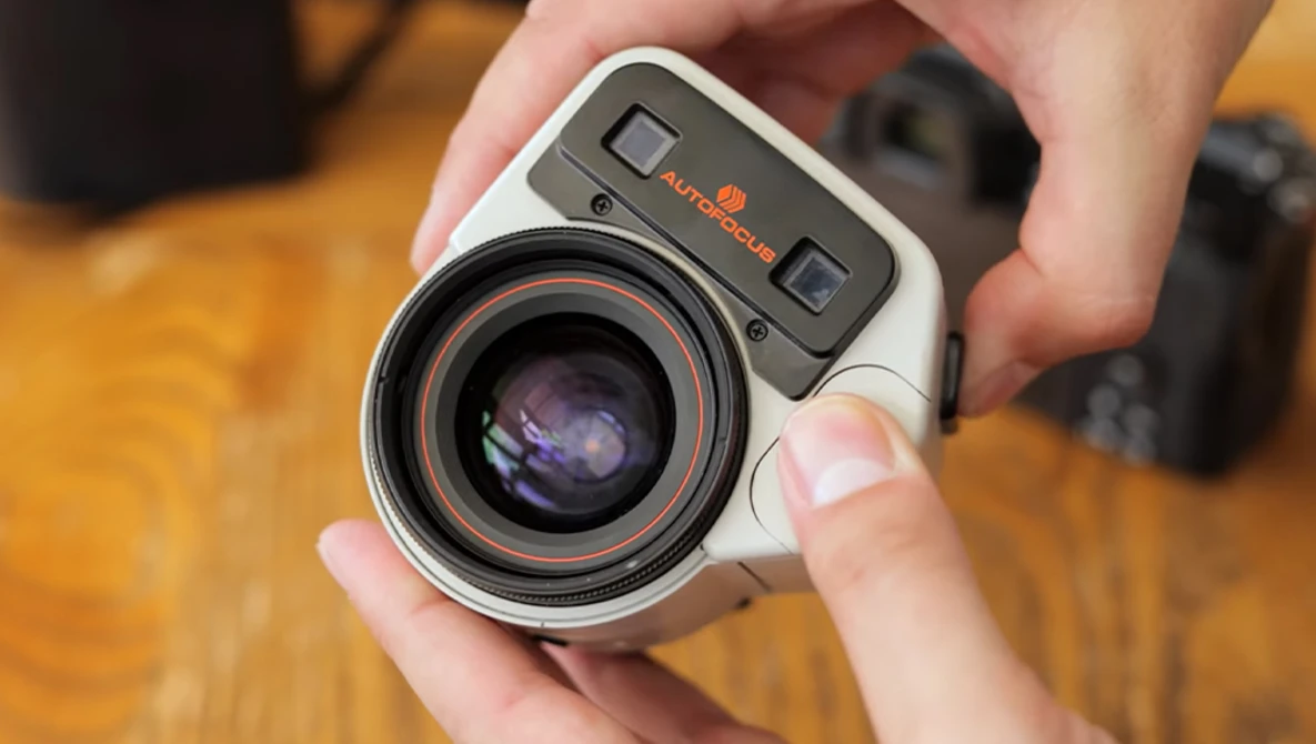Vintage rangefinder camera held in hands, showing lens and top-plate controls against wooden surface.