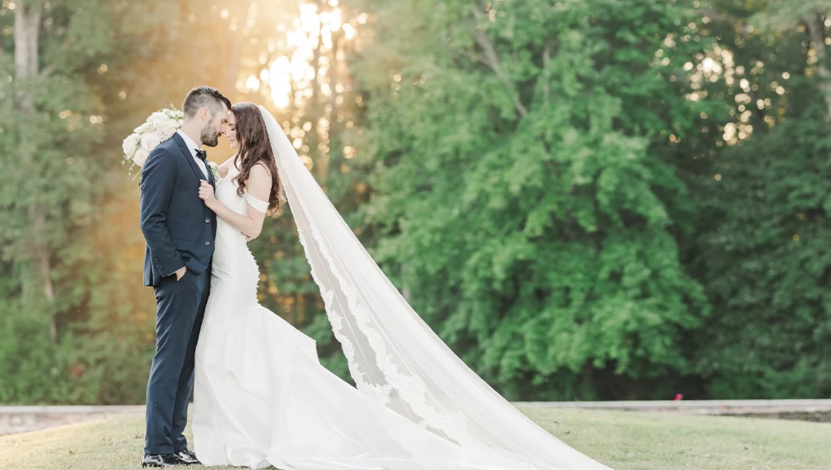 Bride and groom kissing during golden hour with flowing veil and tree-lined background.
