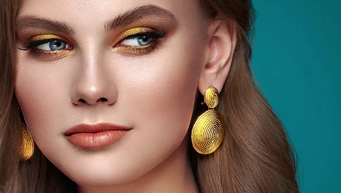 Woman with warm golden eyeshadow and gold teardrop earrings against a teal background.