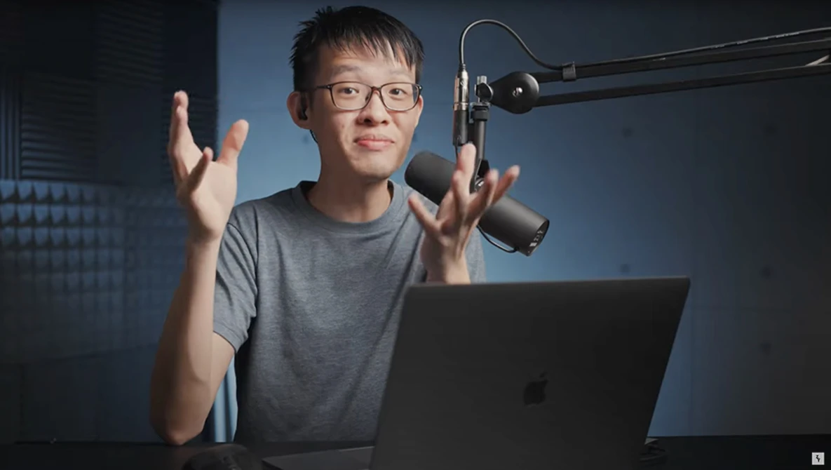 Young person wearing glasses speaking animatedly at a laptop with a studio microphone overhead.