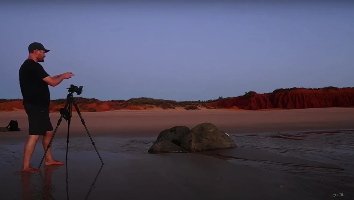 Photographer on a beach at dusk operating a camera on tripod, facing rocky outcrops in shallow water.