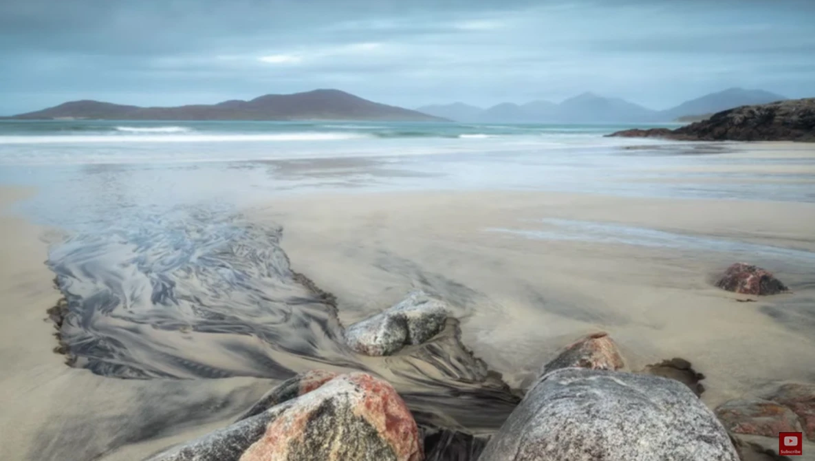 Coastal landscape with sand patterns, rocks, and distant mountains under cloudy sky.