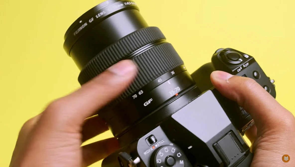 Hands holding a DSLR camera with a telephoto lens mounted against a yellow background.