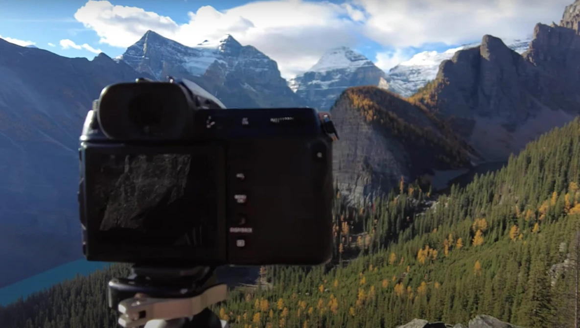 Camera mounted on tripod overlooking alpine valley with snow-capped peaks and forested slopes.