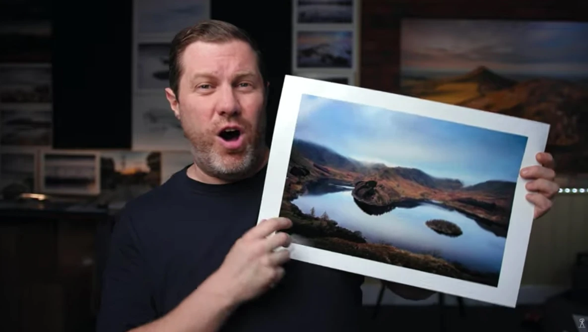 Man holding a framed landscape photograph of a mountain lake with reflective water.