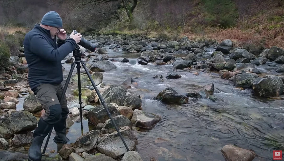 Photographer on tripod shooting by a rocky stream in natural daylight.