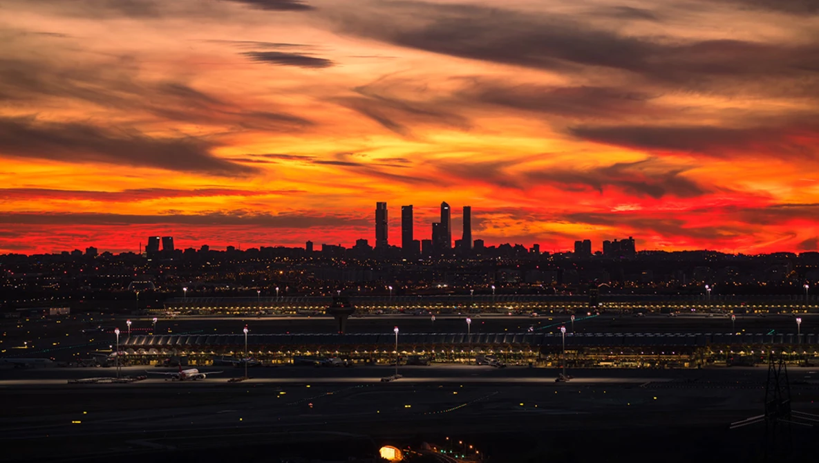 Dramatic sunset over a waterfront city skyline with orange and red clouds illuminating illuminated buildings and a large structure below.