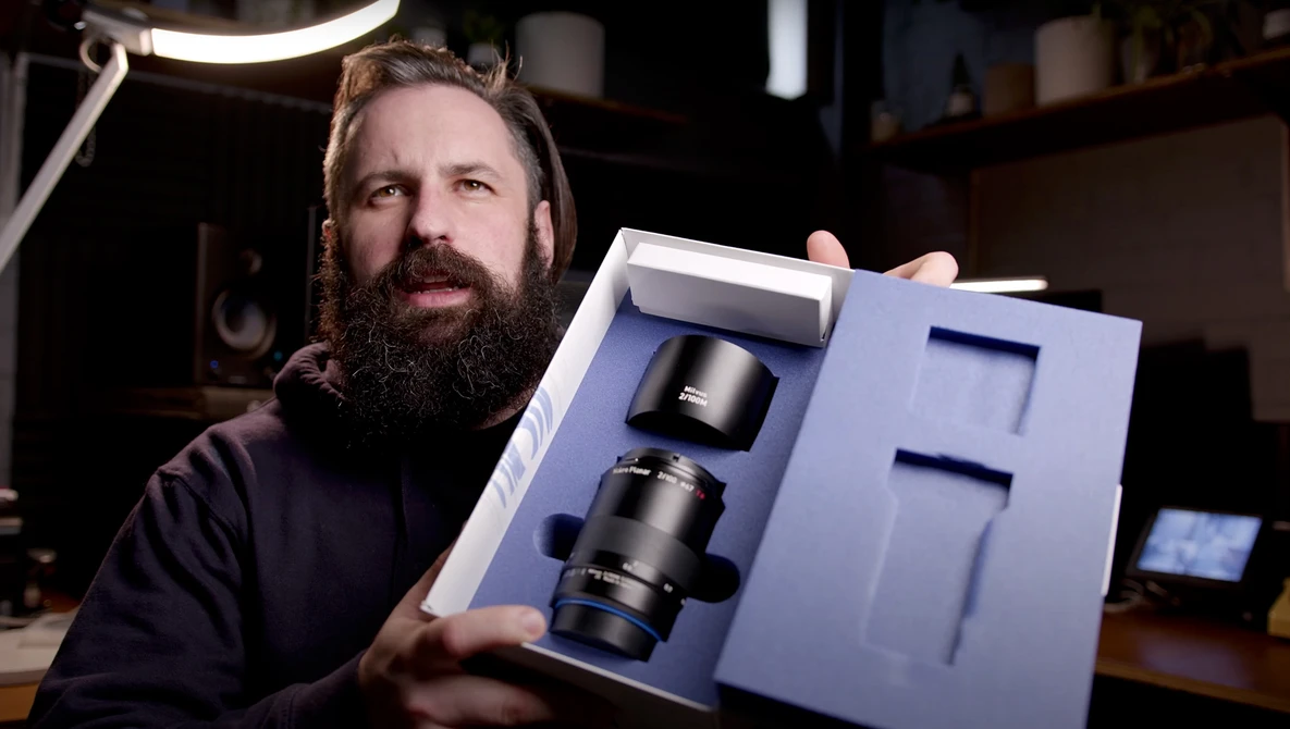 Photographer with beard displaying opened lens packaging containing two camera lenses in a studio setting.