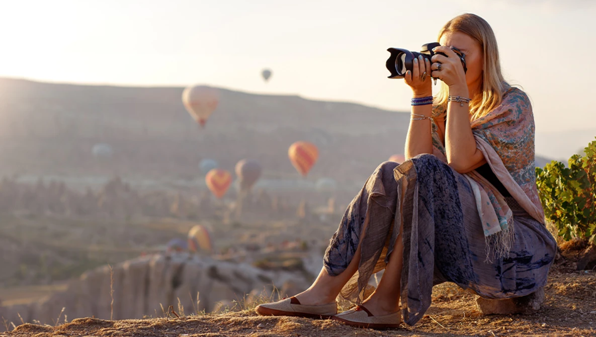 Woman sitting on a hillside photographing hot air balloons floating in a valley at sunrise.