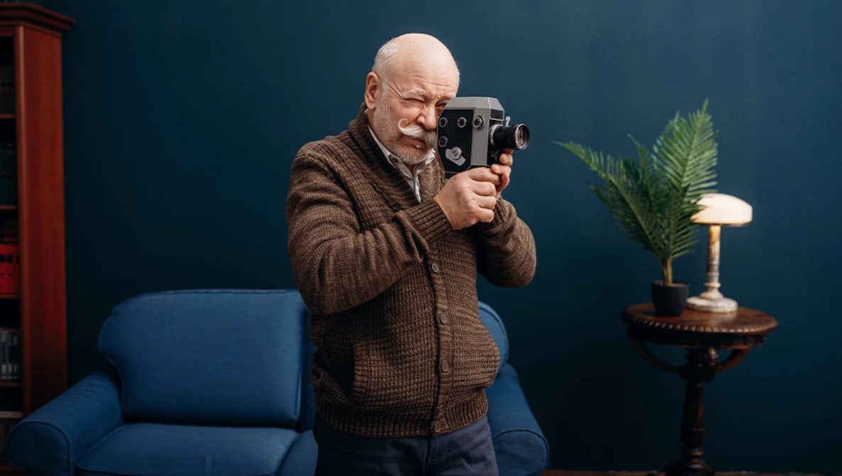 Older man with glasses holding a vintage film camera to his face while standing indoors.