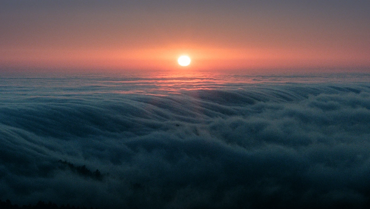 Sunrise over a sea of clouds with the sun glowing orange above the cloud layer.