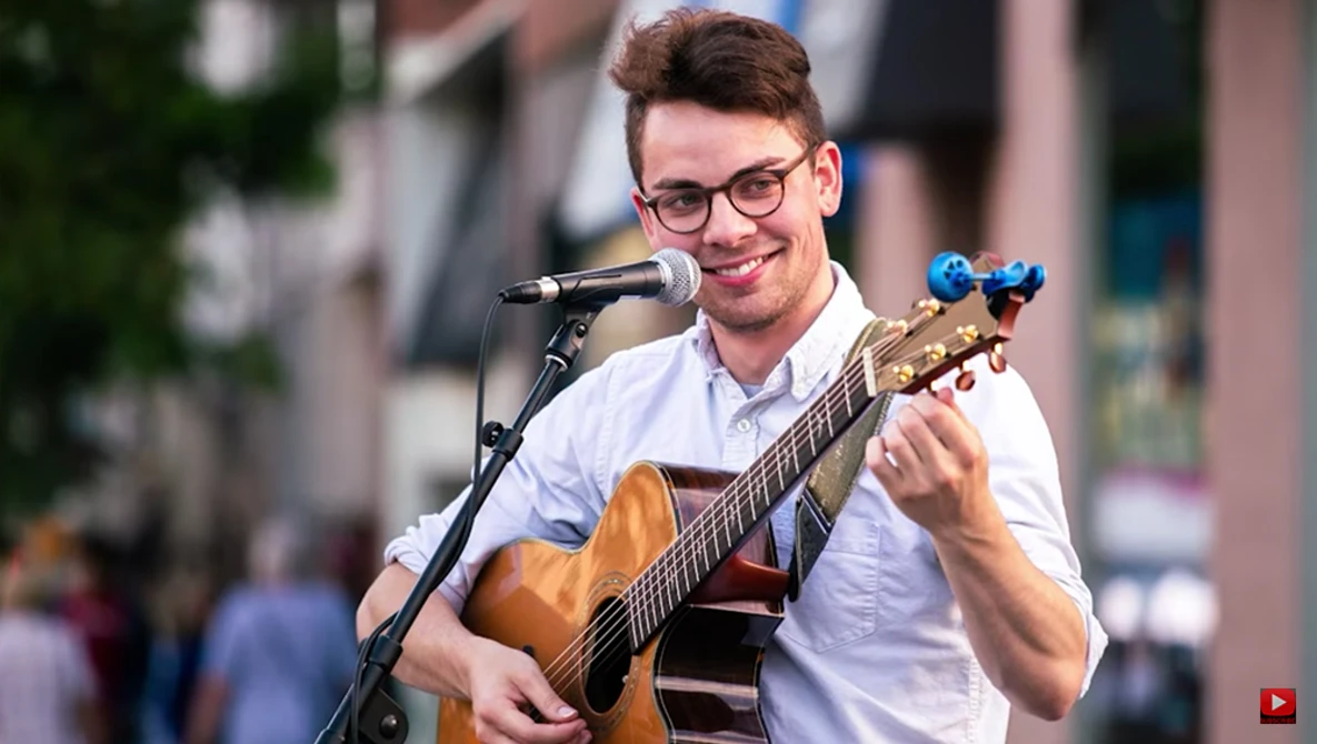 Musician performing outdoors with acoustic guitar and microphone, wearing glasses and light blue shirt.