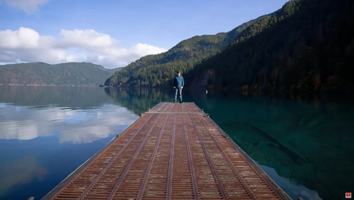 Person standing at the end of a wooden pier extending into a calm alpine lake surrounded by forested mountains.