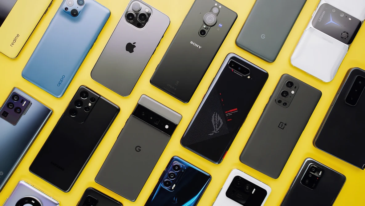 Flat lay of approximately 12 smartphones arranged on a bright yellow background, displaying various models and finishes.