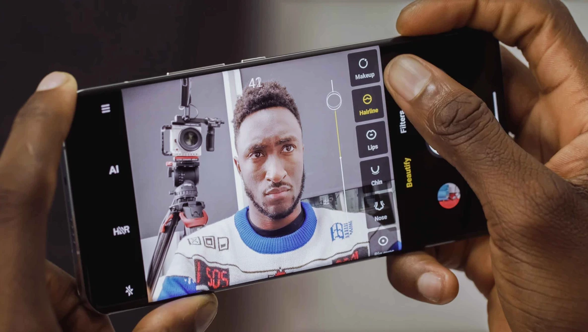 Smartphone camera interface displaying a portrait of a person with a camera mounted on a tripod visible in the mirror reflection.