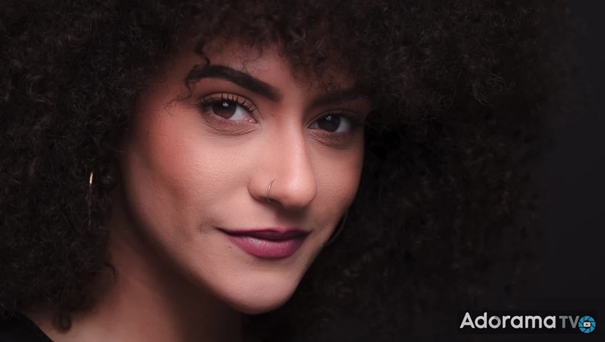 Portrait of a woman with curly dark hair against a dark background, lit with warm side lighting.