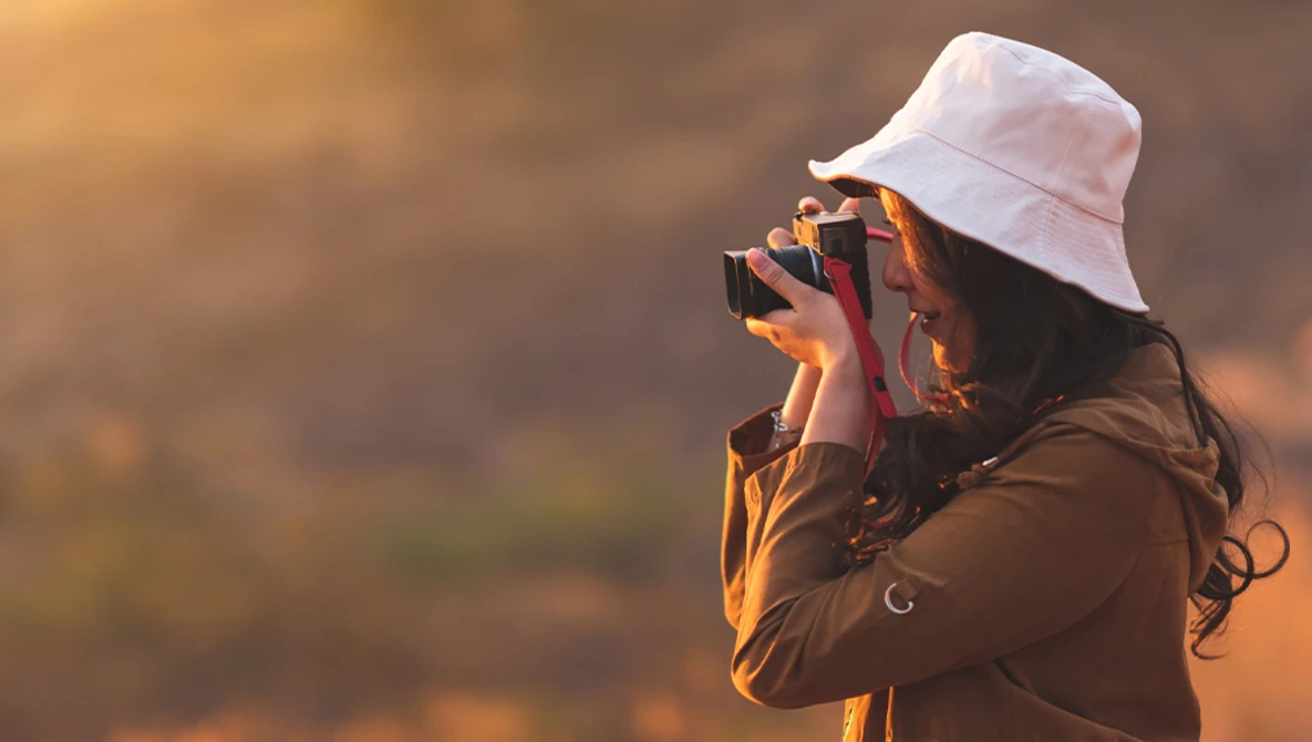 Photographer in white bucket hat holding camera to face, backlit by golden hour sunlight.