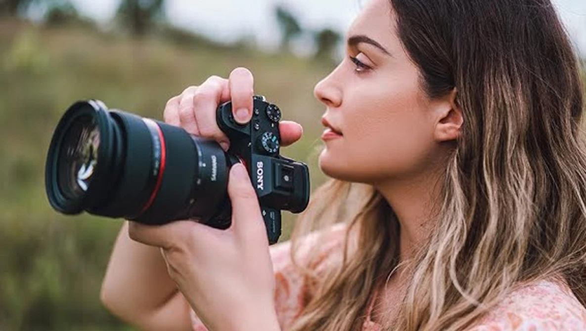 Woman holding a mirrorless camera with telephoto lens to her eye while focusing on a distant subject.