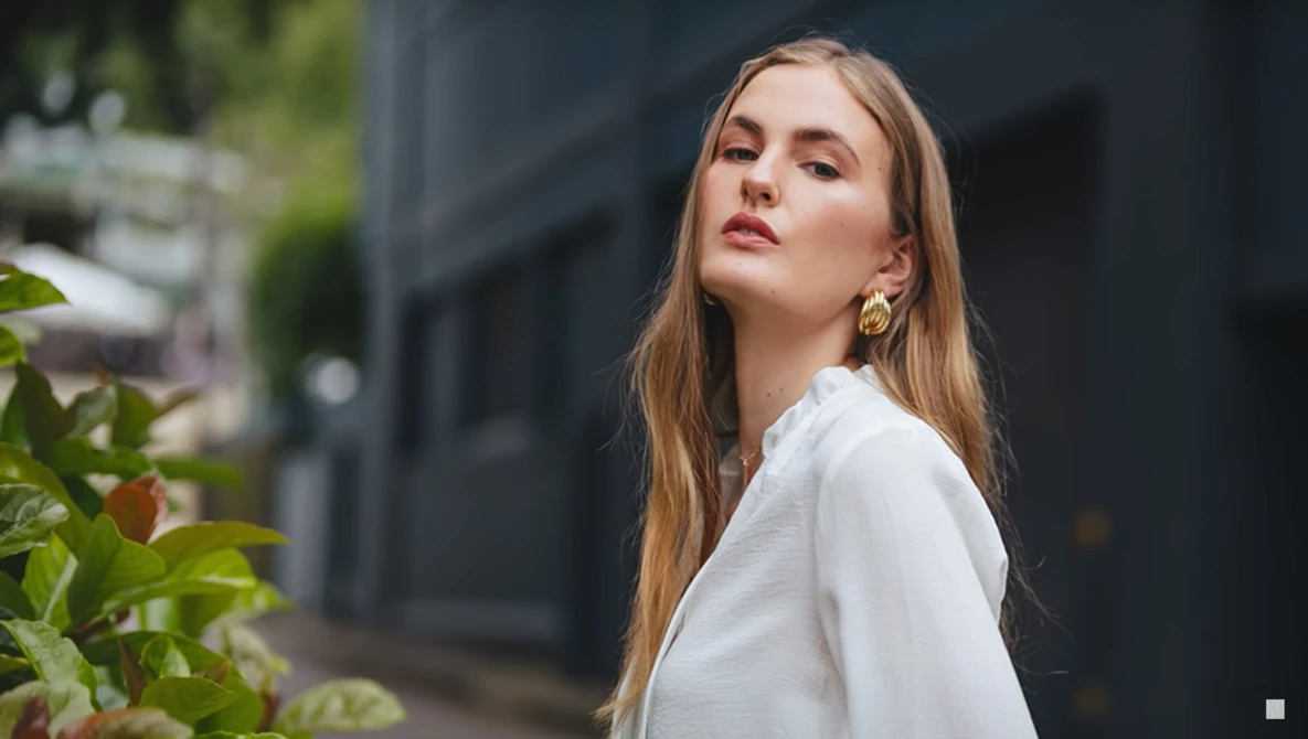 Woman with long reddish-blonde hair wearing a white button-up shirt, looking upward against a blurred dark building backdrop.