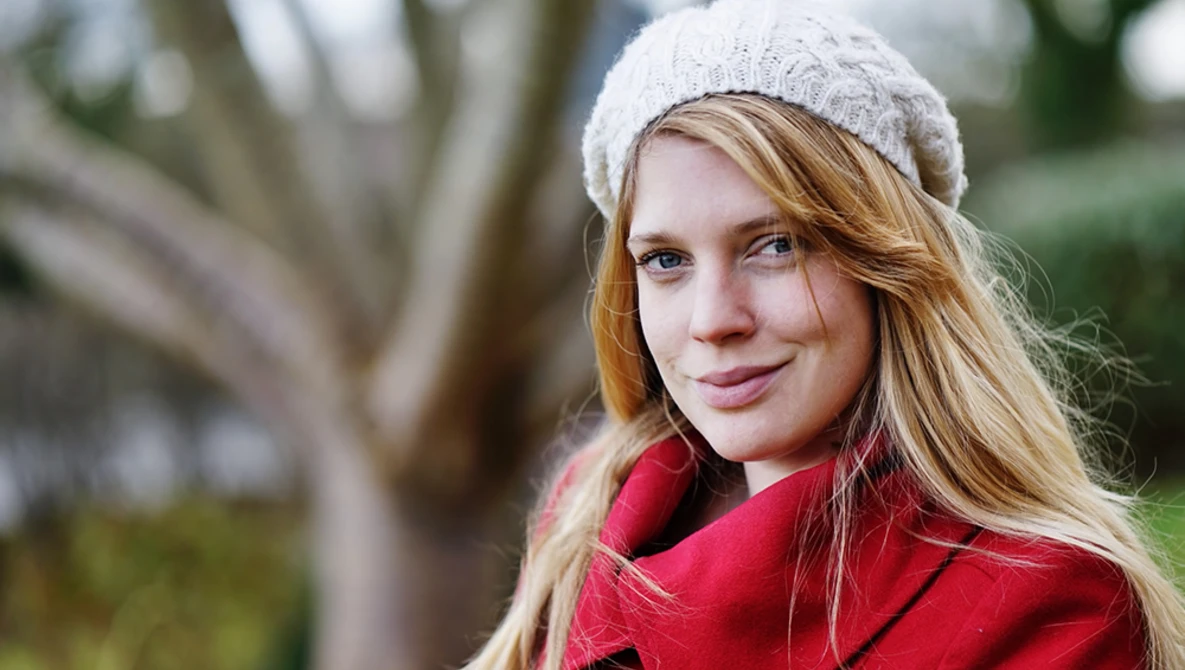 Young woman with long reddish-blonde hair wearing a white knit beanie and red jacket, smiling at camera outdoors.