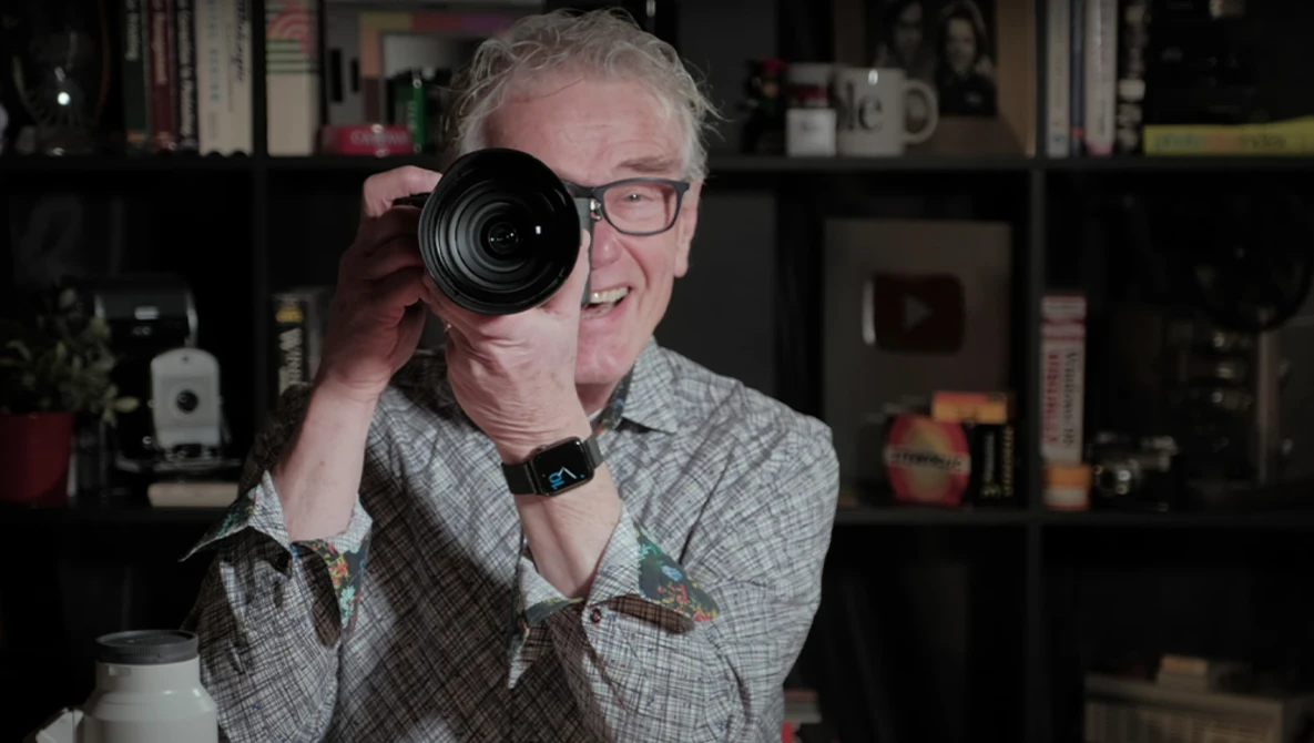Photographer holding a telephoto lens up to his eye, smiling at the camera in a home studio setting.