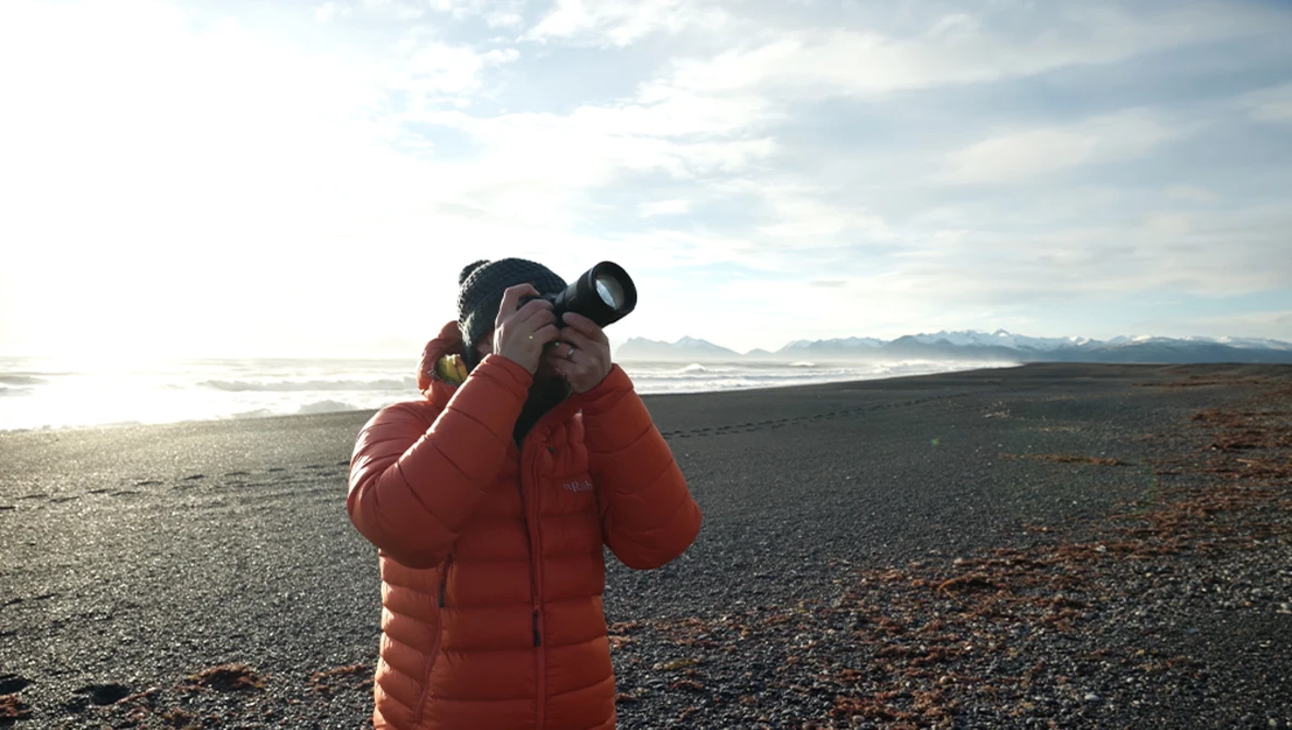 Photographer in red jacket holding binoculars to face while standing on dark volcanic terrain with mountains visible in distant background.