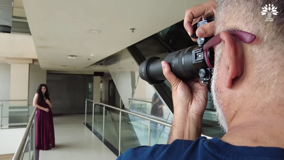 Photographer holding a mirrorless camera with telephoto lens, shooting a woman in a burgundy dress posed on a modern indoor balcony.
