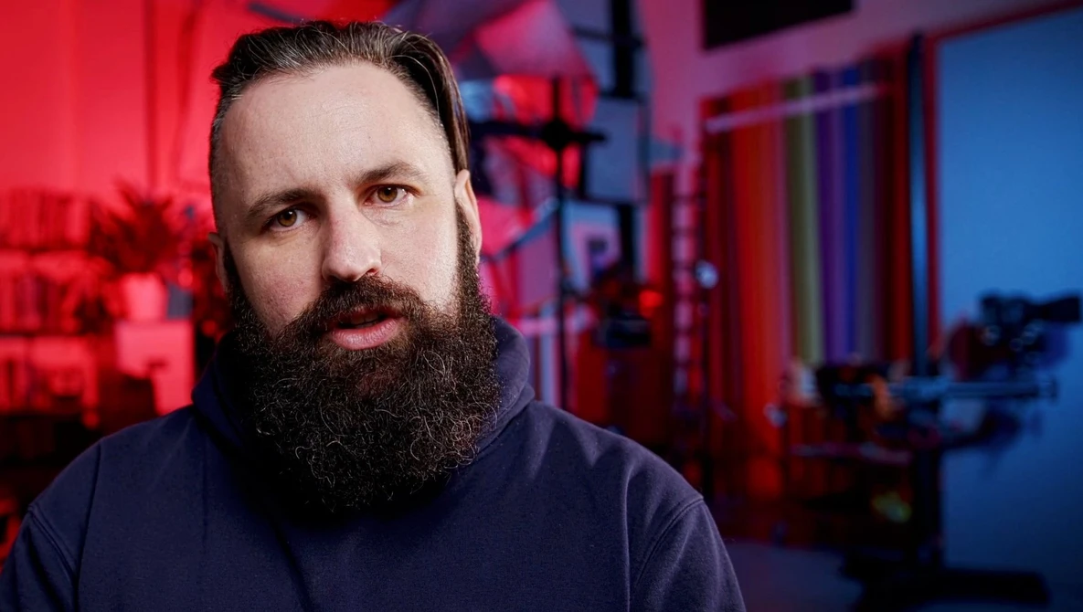 Bearded man with dark hair in a dark hoodie, photographed against red and blue neon lighting.