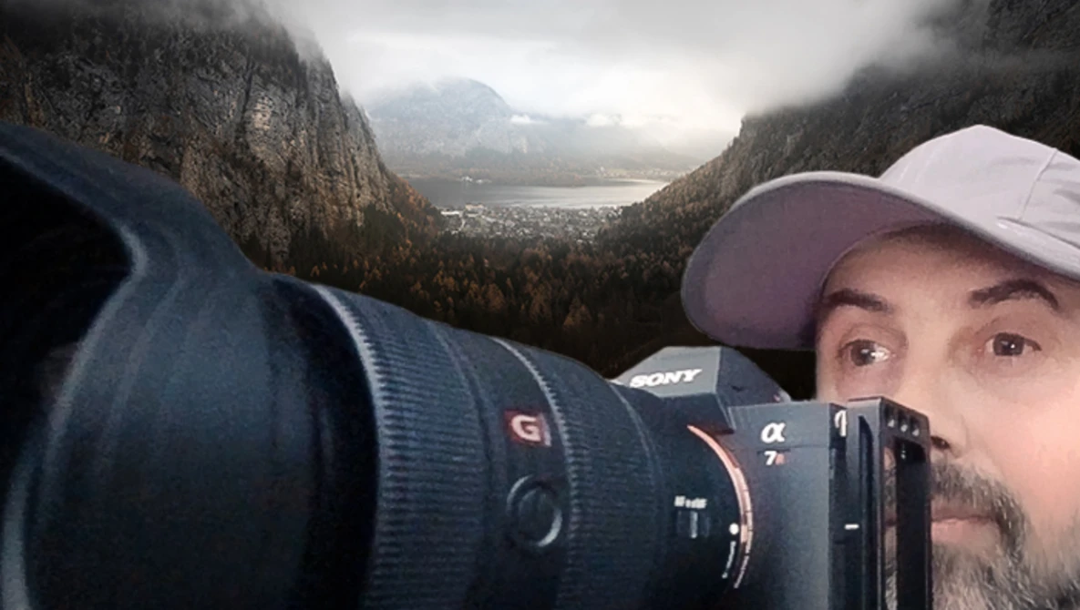 Photographer holding Sony mirrorless camera with telephoto lens, smiling at camera with misty alpine lake and forested mountains in background.