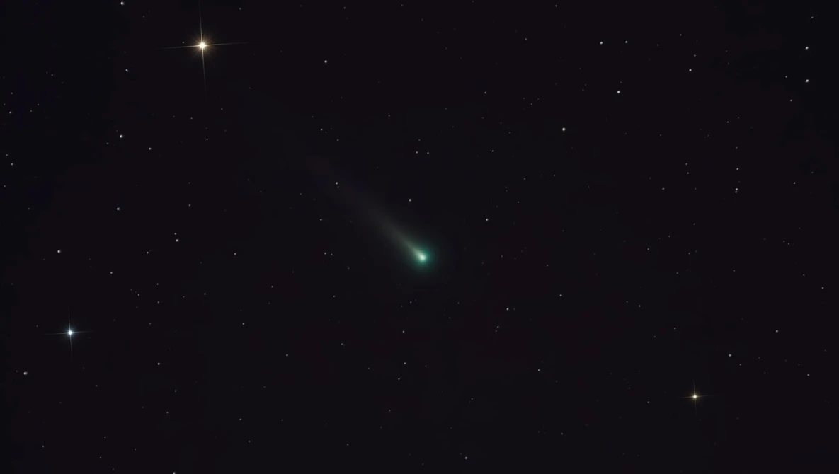 Comet with greenish coma and tail streaking across dark starfield at night.
