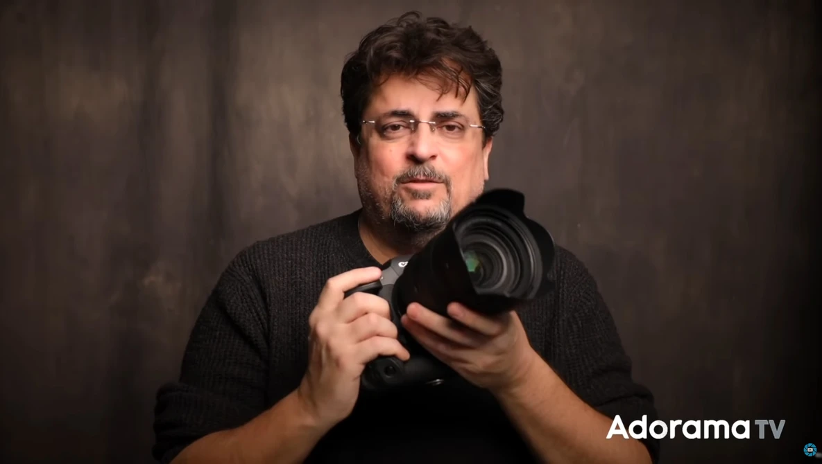 Man holding a camera lens toward the viewer against a neutral backdrop.