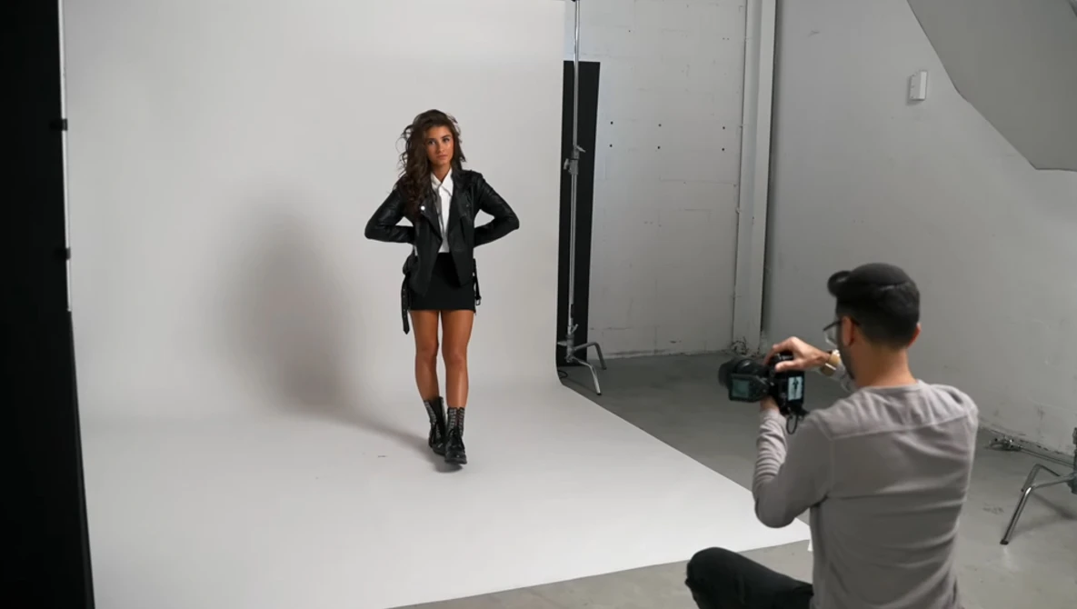 Photographer shooting a model in a studio with white backdrop and professional lighting setup.