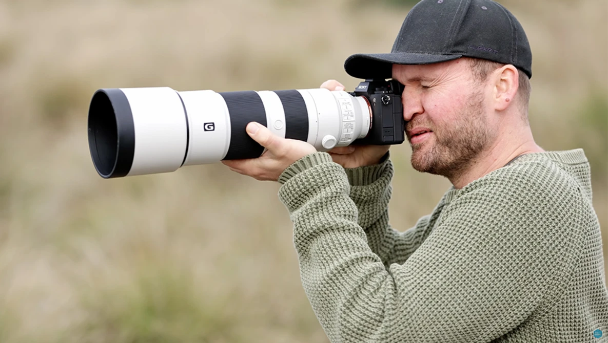 Photographer with black cap holding a telephoto lens with white barrel and black accents, looking through the viewfinder.