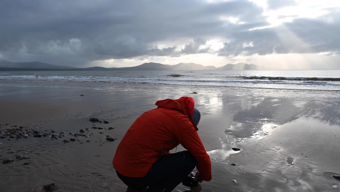 Person in red jacket crouching on wet sand at beach with mountains in background under dramatic cloudy sky.