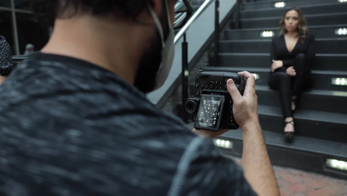 Photographer holding a mirrorless camera with articulating screen, capturing a portrait of a woman posed on illuminated stairs.