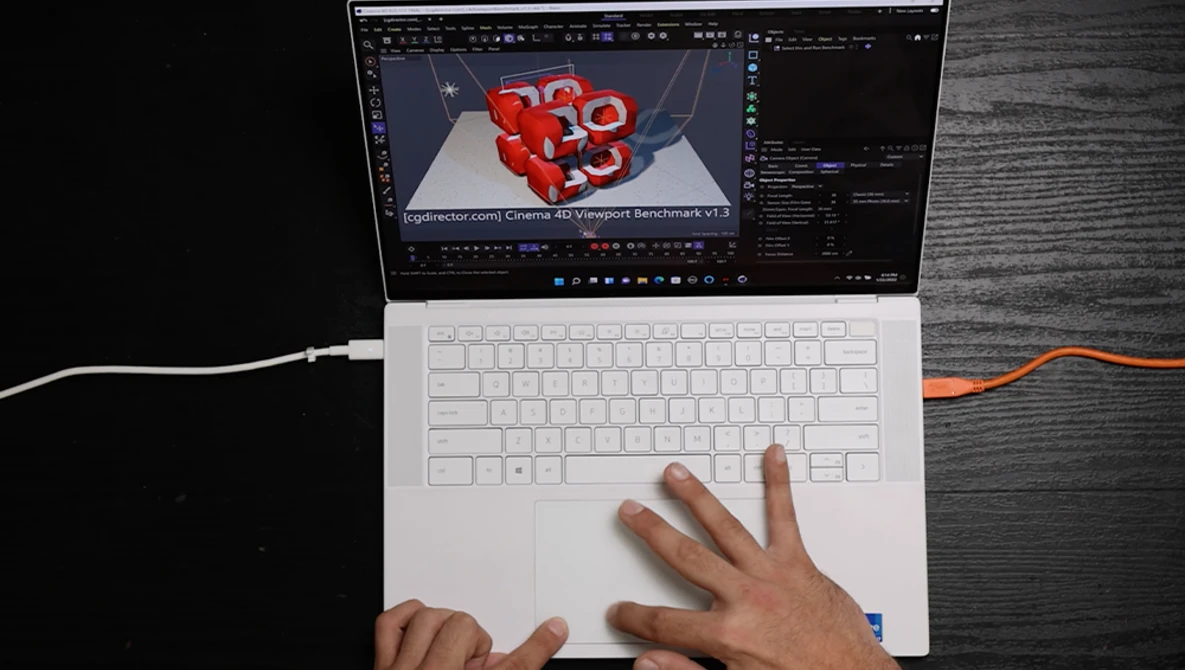 Overhead view of hands on a white laptop trackpad while 3D design software with red geometric shapes displays on screen.