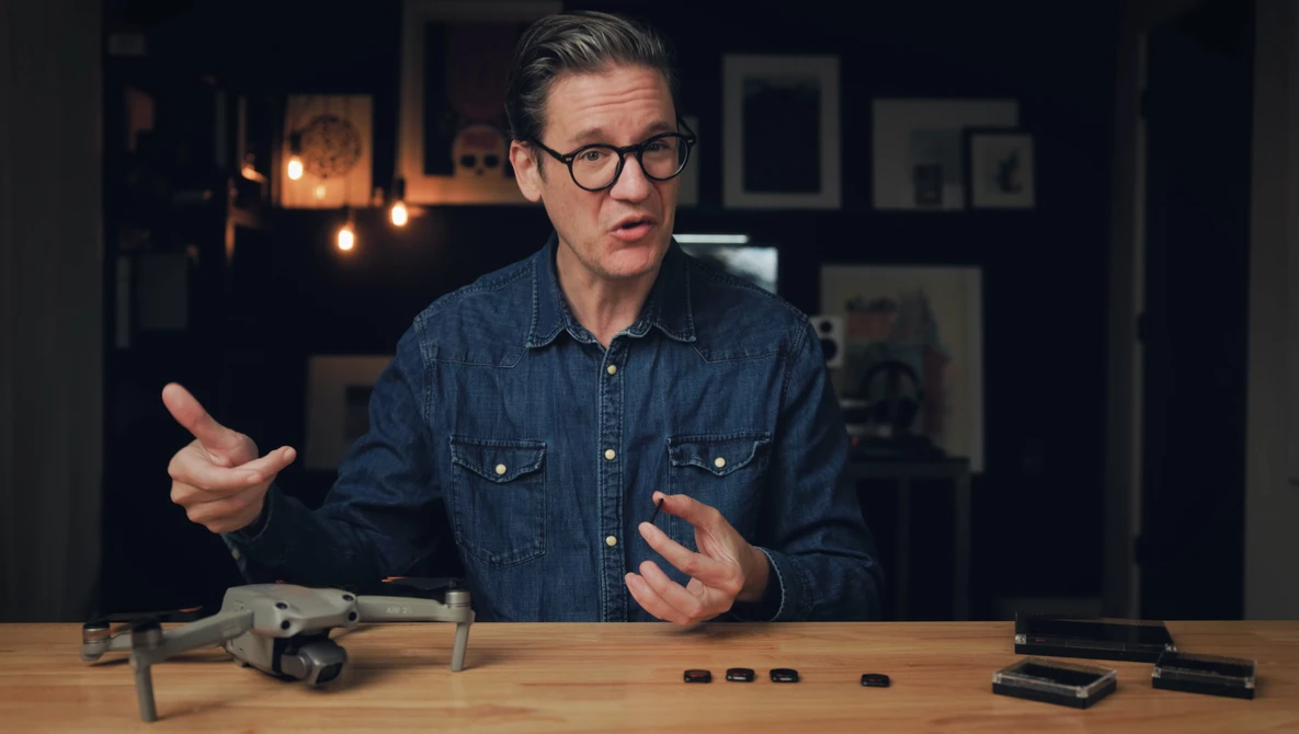 Man in denim shirt gesturing while discussing camera equipment on wooden table.
