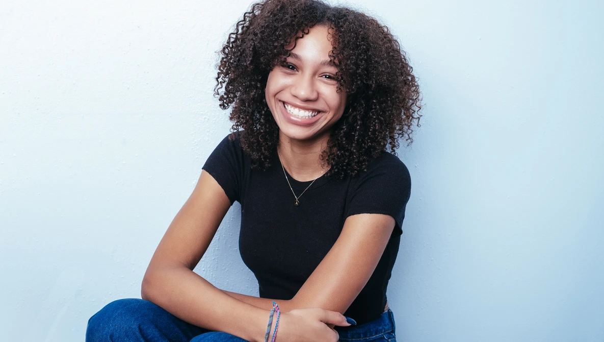 Woman with curly dark hair smiling at camera while seated against light blue background.