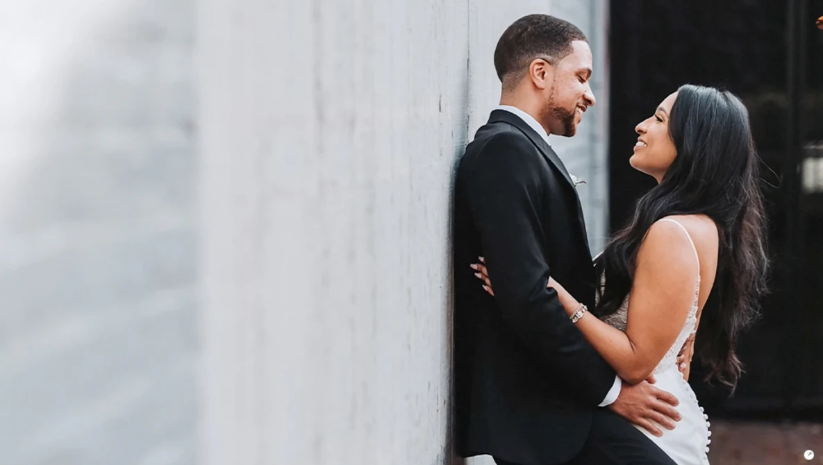 Couple in formal attire leaning against a white brick wall, looking at each other intimately.