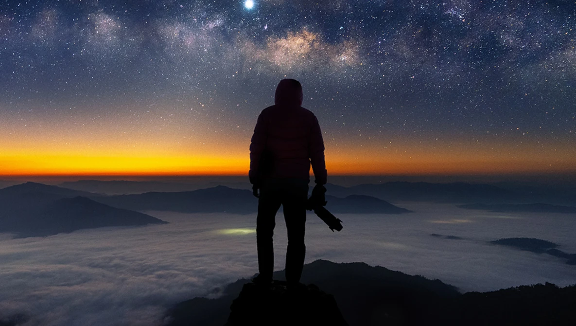 Silhouetted figure standing on a mountain peak overlooking a sea of clouds at sunrise, with the Milky Way visible in the night sky above.