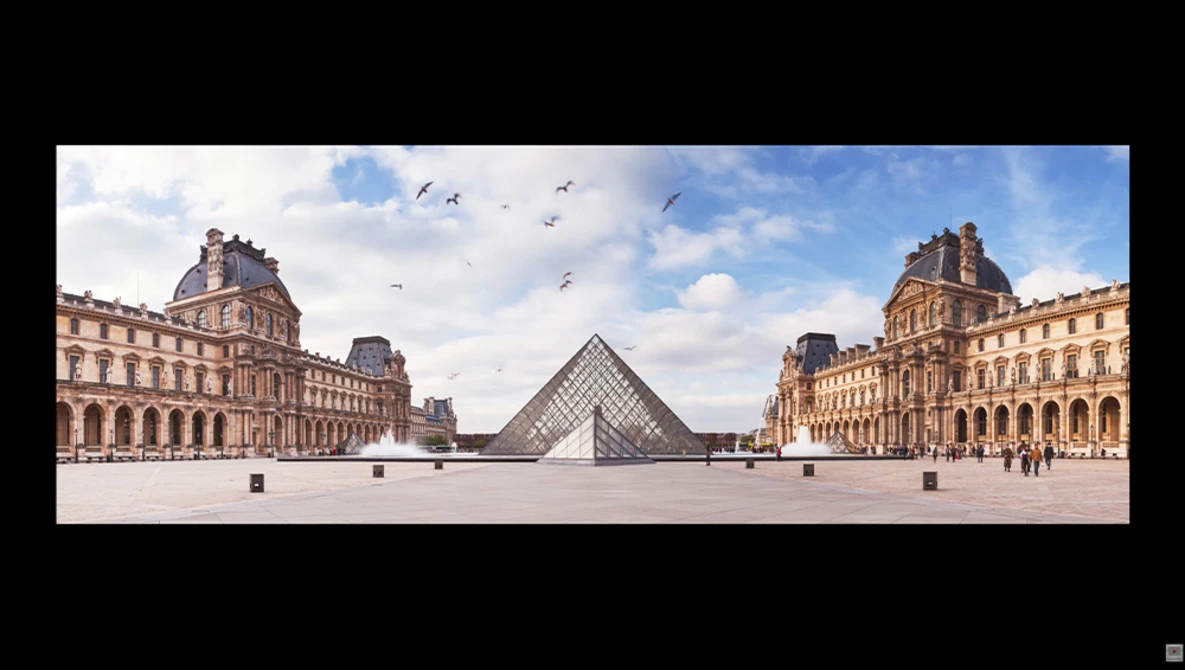 Glass pyramid structure centered in a vast courtyard surrounded by classical palace architecture under blue sky.