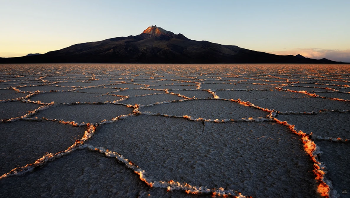 Volcanic landscape at dawn with cracked earth patterns and a cone-shaped volcano silhouetted against the sky.