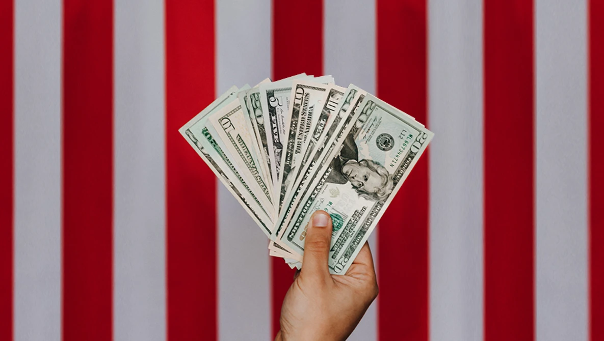 Hand holding a fan of U.S. dollar bills against a red and white striped background.