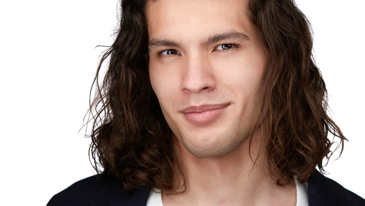 Man with long dark wavy hair wearing a dark blazer over a white shirt against a white background.