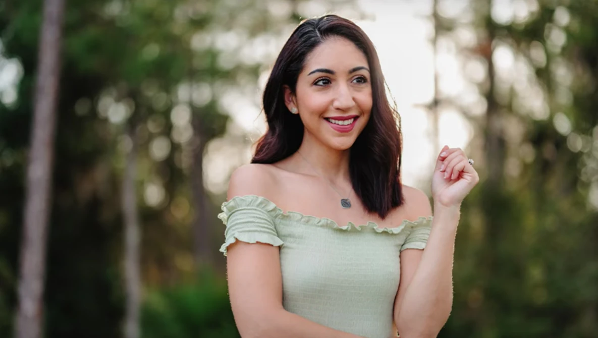 Woman with long dark hair wearing an off-shoulder sage green top, smiling outdoors among tall trees.