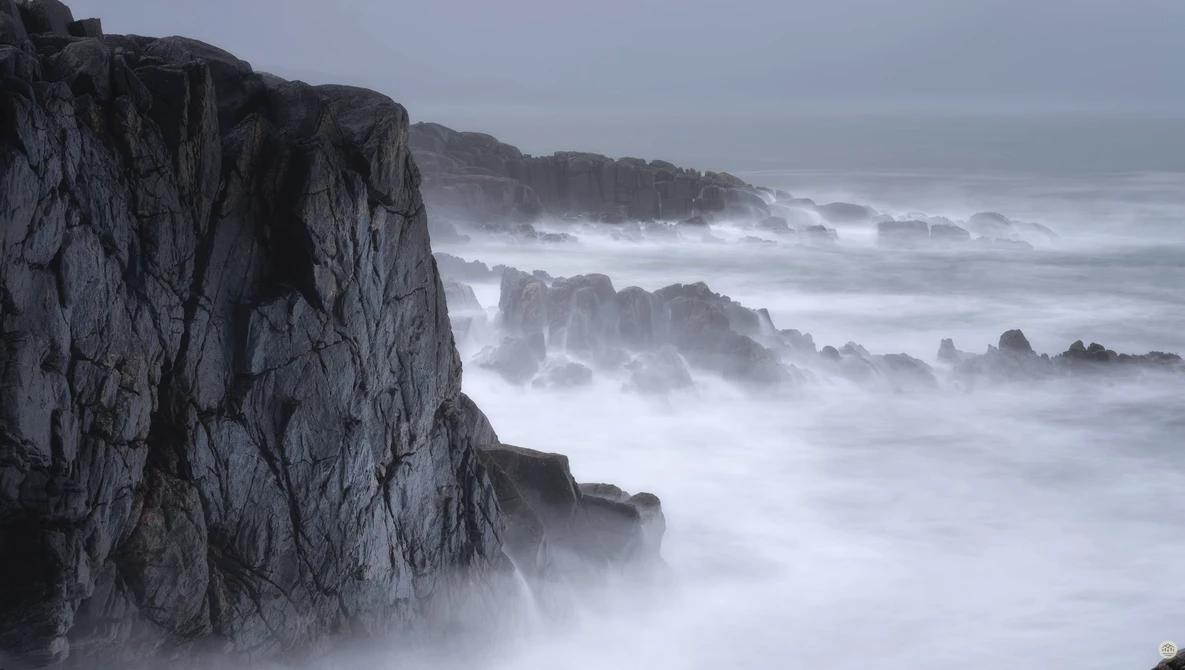 Dramatic rocky coastline with crashing waves and mist shrouding distant cliffs.