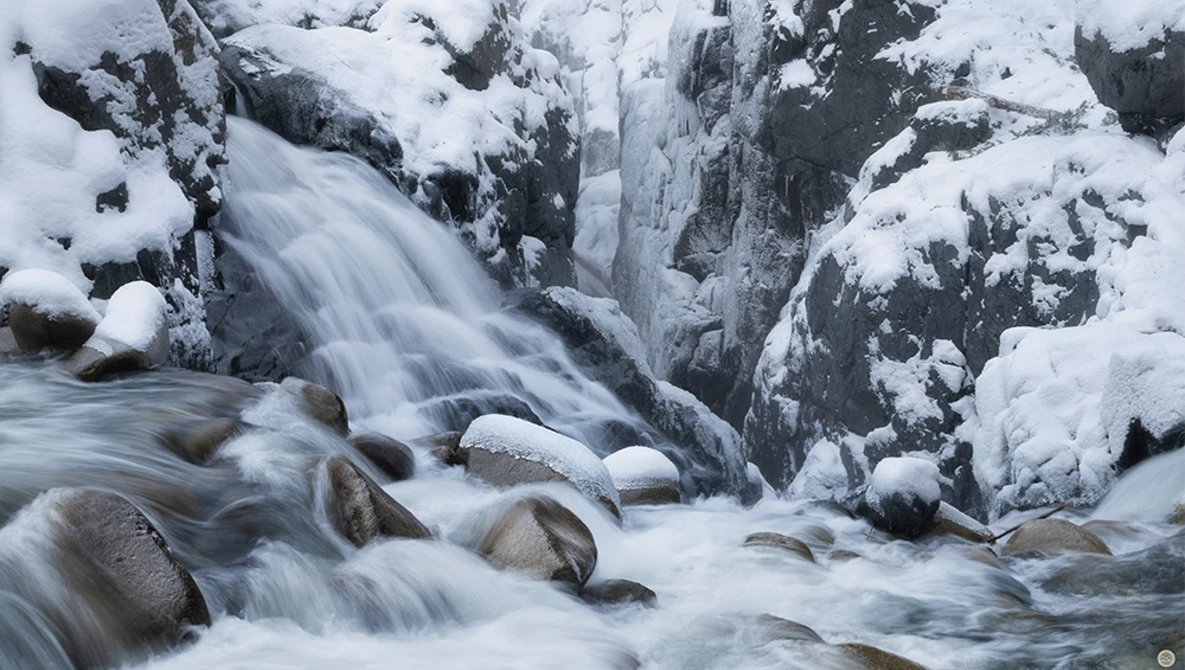 Frozen waterfall cascading over snow-covered rocks and boulders in a winter landscape.
