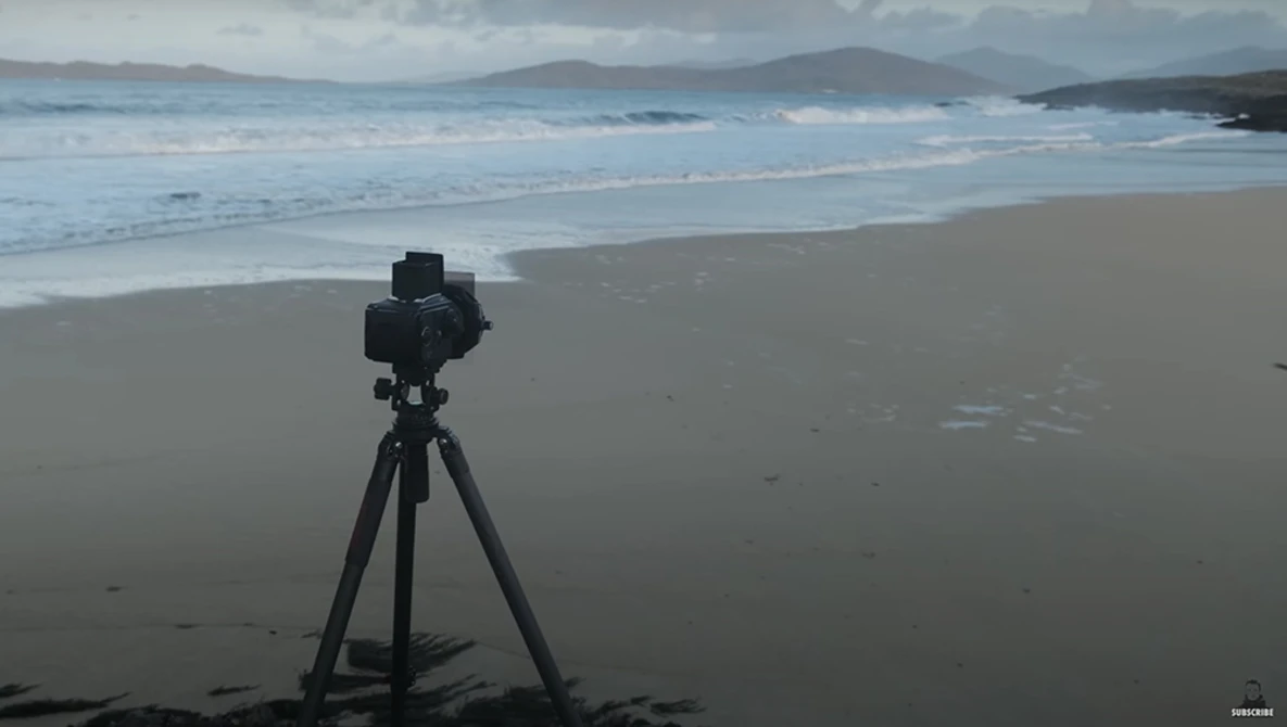 Camera on tripod positioned on a beach facing the ocean and distant mountains.