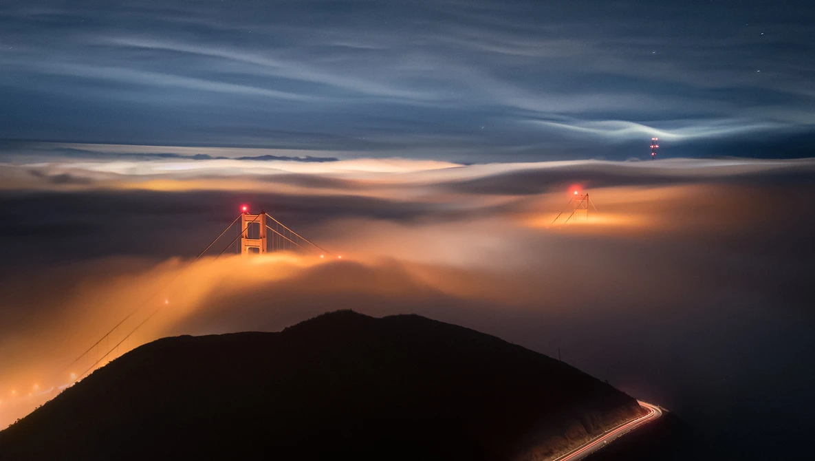 Golden Gate Bridge emerging from thick fog at twilight, illuminated by warm sunset light above a dark hillside silhouette.